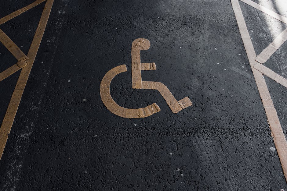 A parking space marked with a white wheelchair accessibility symbol painted on the dark asphalt surface, located outdoors near a building entrance. The marking is centrally positioned within the space, with a wooden border outline visible around the symbol. To the left, part of a yellow-striped chair or barrier is seen, while on the right, there are additional parking lines and a paving edge. The environment appears to be well-lit, indicating daytime, and the area is designated for disabled parking as part of the home relocation or furniture transport logistics managed by Man with Van Watford, supporting efficient access and unloading during moving services at Cassiobury Park.